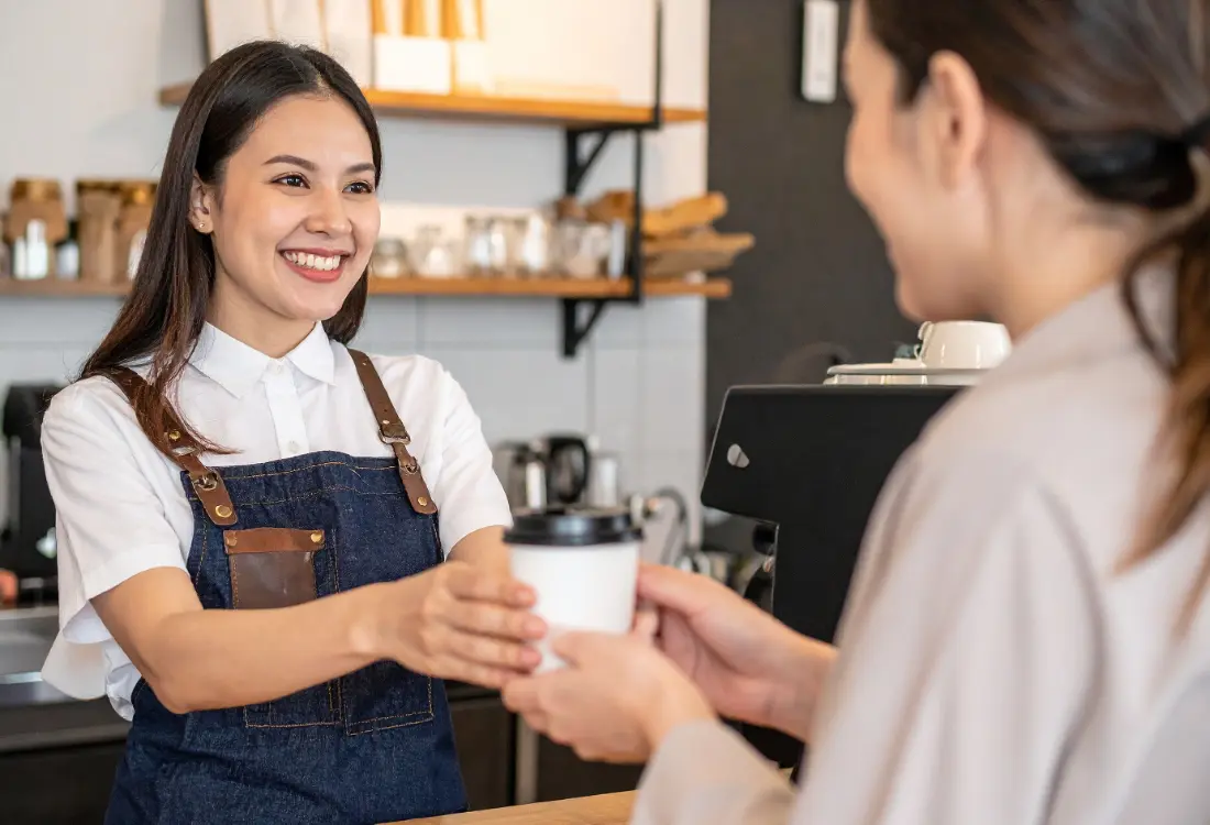 Barista wearing a collared work uniform polo shirt with embroidered logo, offering a coffee cup to a client in a professional workplace setting.