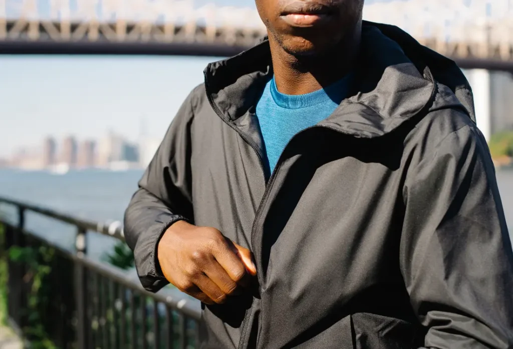 Man wearing a lightweight hooded jacket over a blue T-shirt by the waterfront, illustrating how outdoor-inspired clothing is worn for walking and everyday urban activities.