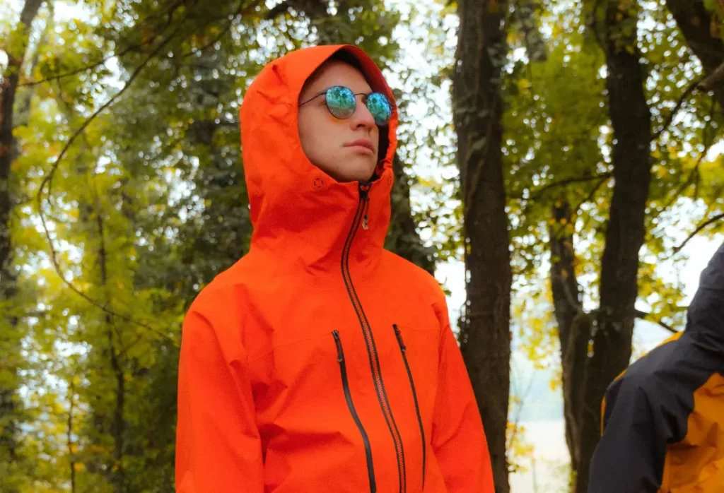 Bright orange gorpcore jacket with a hood, front zips and reflective sunglasses, showing the functional outdoor gear that people love for its bold colours and practical design.